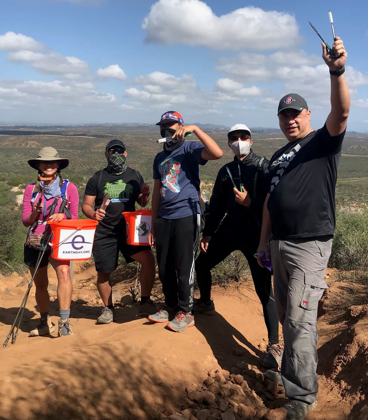 Volunteers stand on a dirt trail holding trash buckets.
